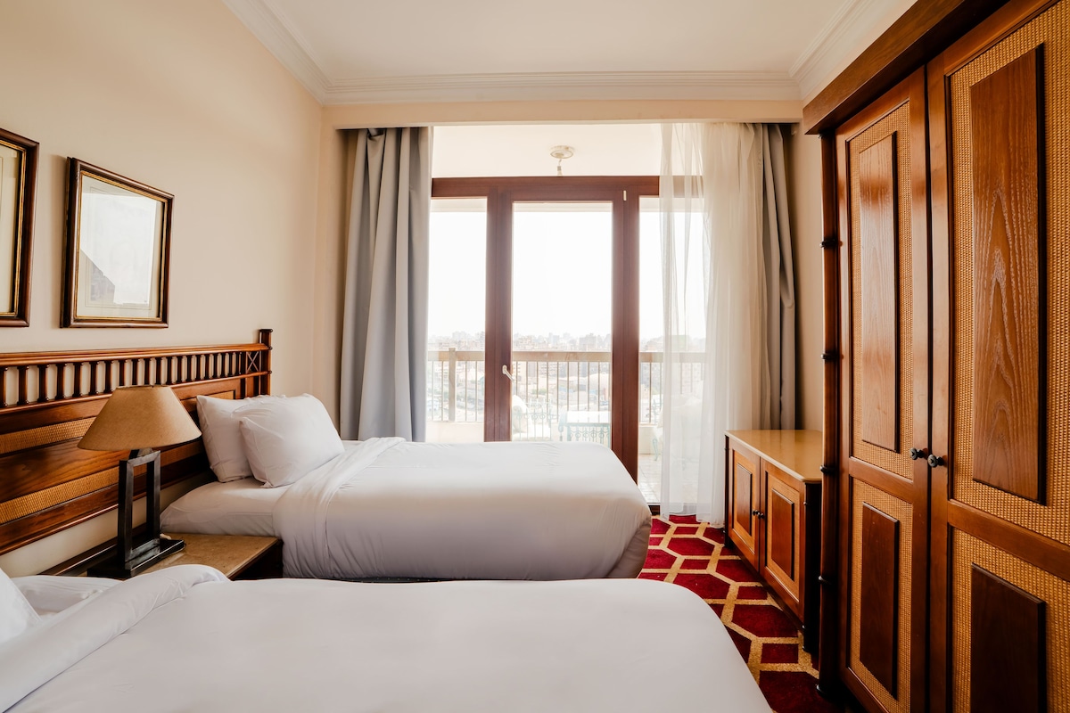 A hotel room with two beds, a wooden wardrobe, a desk with a lamp, and a view of the city through a large window.
