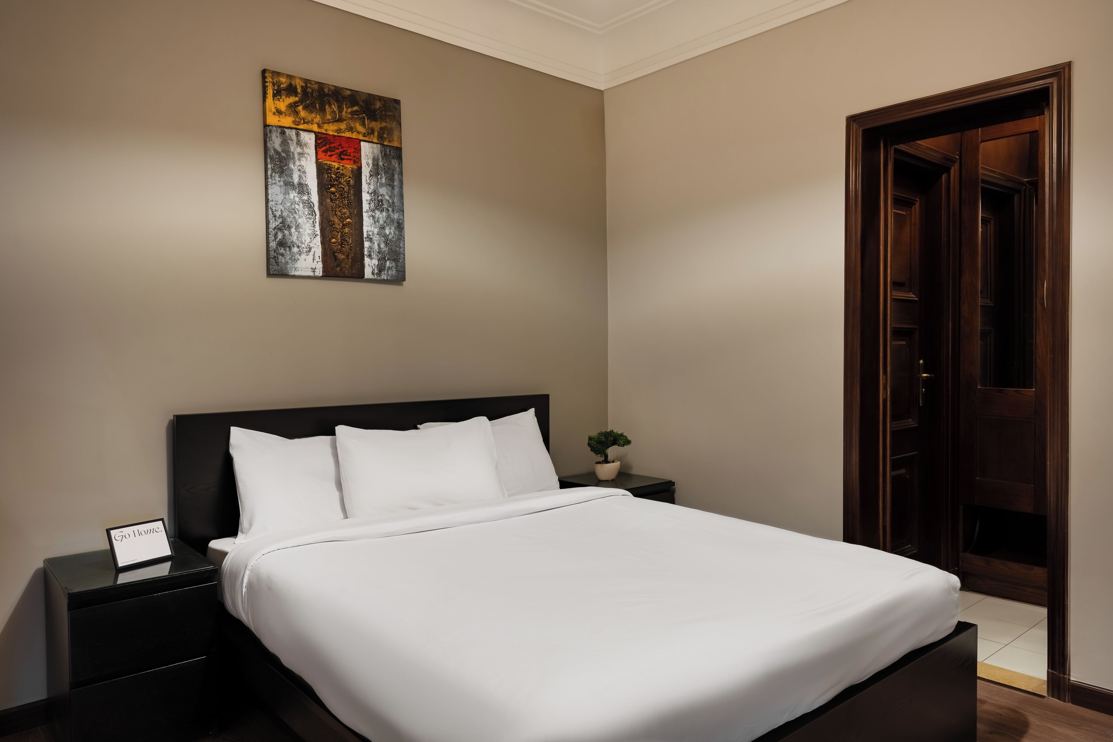Una cama bien hecha en una habitación de hotel, con un cabecero oscuro, una mesita de noche pequeña con una lámpara y una obra de arte colgada en la pared.