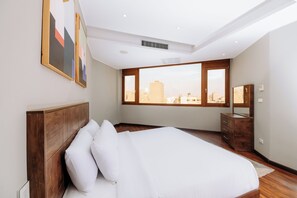 Een slaapkamer met een groot bed, een houten dressoir en een stadszicht door grote ramen.