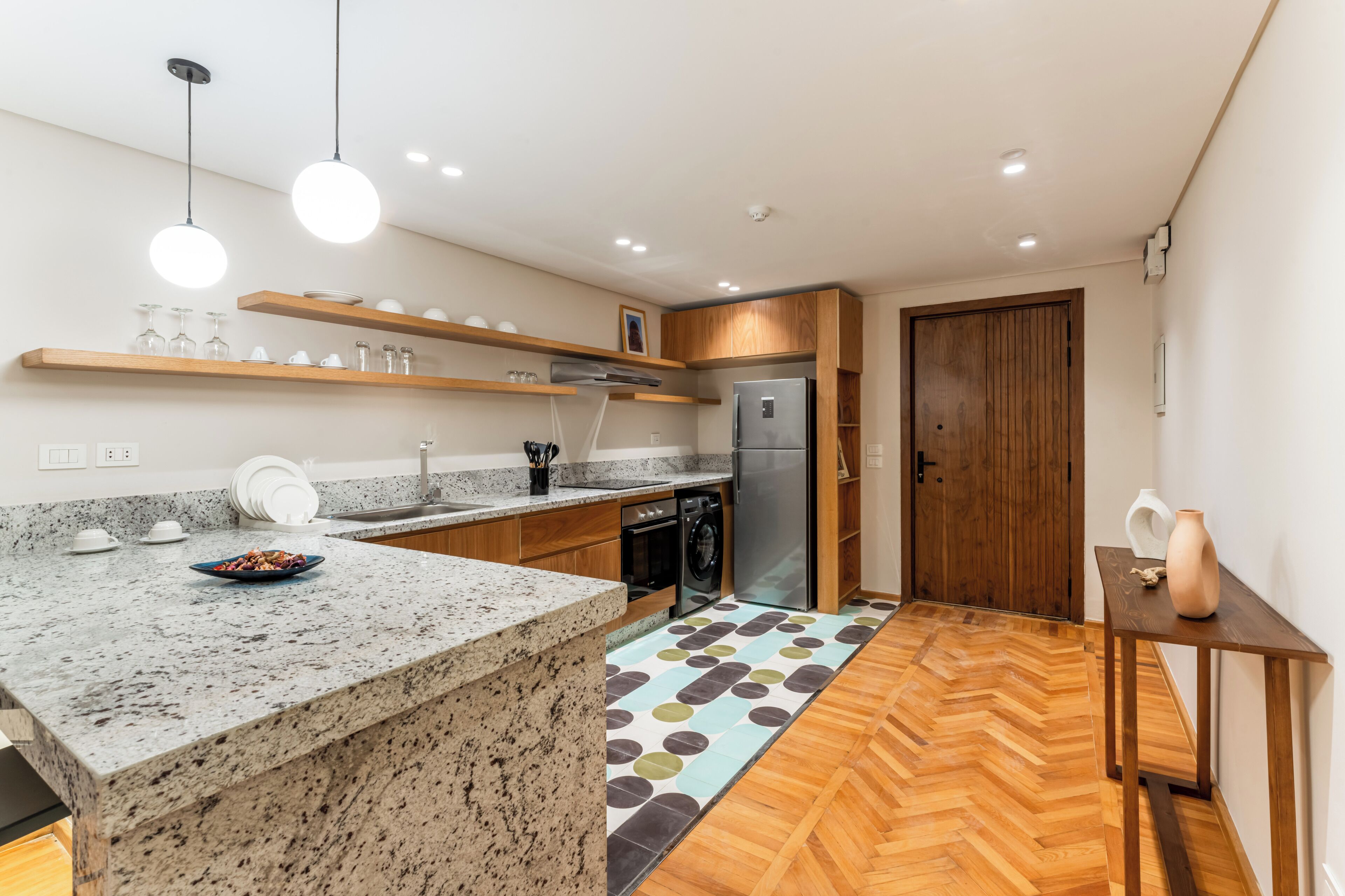 A modern kitchen with granite countertops, wooden shelves, a built-in oven, and a refrigerator.