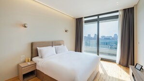 A modern hotel room with a large bed, a nightstand with a phone and candles, a view of the city through large windows, and a cityscape visible outside. - Brassbell Zamalek Om Kolthoom (Cairo)