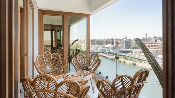 A balcony with wicker chairs and a glass door leading to an interior space. A river and cityscape are visible in the background. - Brassbell Zamalek Om Kolthoom (Cairo)
