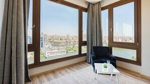 A spacious room with large windows, a dark blue chair, and a small table with a glass and a green object. - Brassbell Zamalek Om Kolthoom (Cairo)
