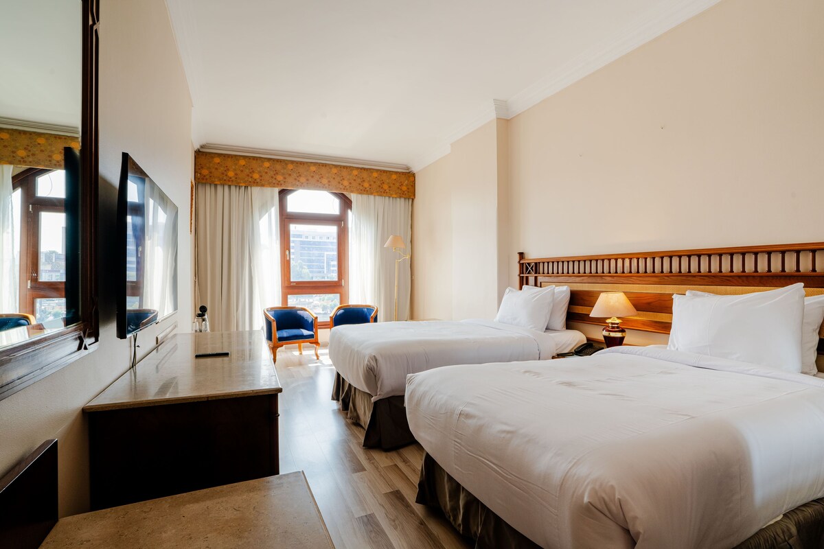 A hotel room with two beds, a desk, a chair, a lamp, and a large mirror.