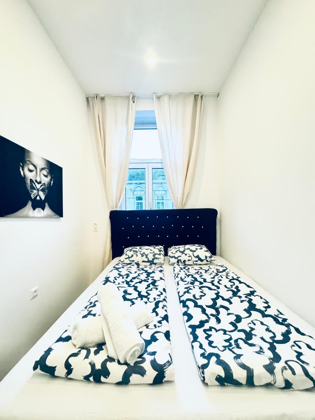 A bedroom with a bed, curtains, a painting, and a window with a view of buildings.