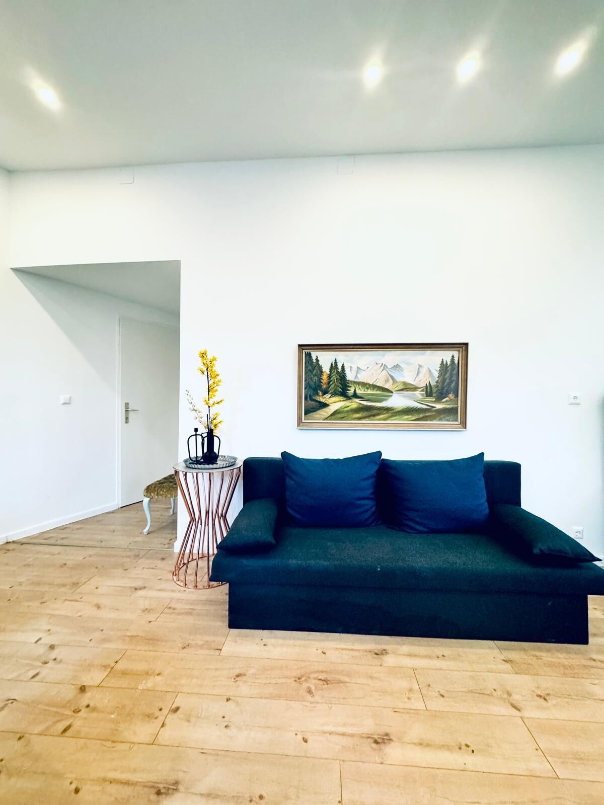 Un salon moderne avec un canapé vert foncé, un tableau représentant un paysage encadré et une petite table d’appoint sur laquelle est posé un vase rempli de fleurs jaunes.