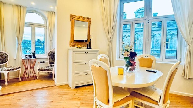 A dining area with a round table, white chairs, and a vase with flowers. There is a window with curtains and a view of buildings outside. - Comfortable 3BR Stay Kaufpark Area (Vienna)