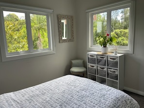 Quarto com cama, duas janelas com vista para o verde, uma cadeira pequena e um móvel branco com gavetas.