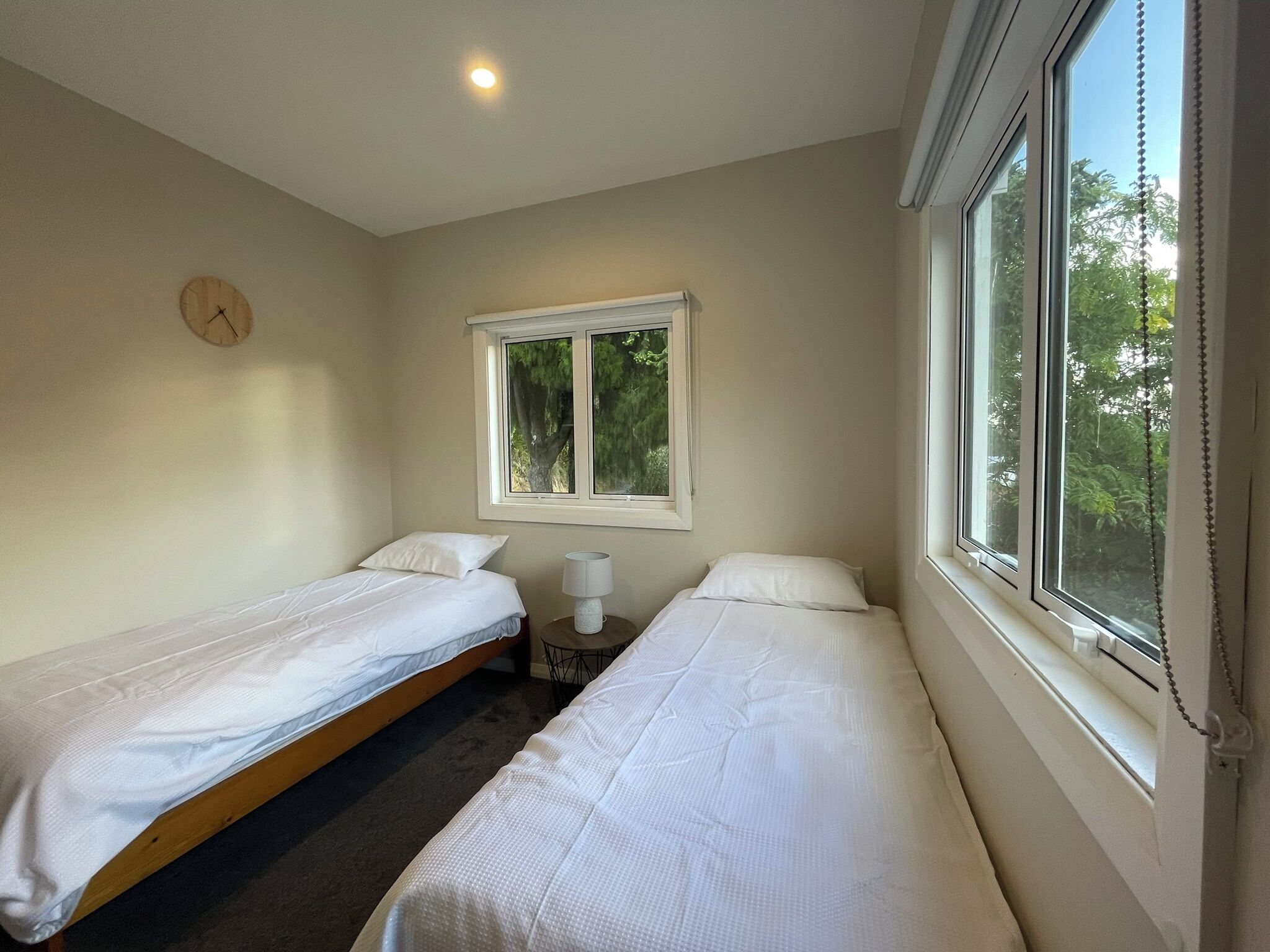 A room with two single beds, a small table, a lamp, a clock on the wall, and a window with a view of trees.