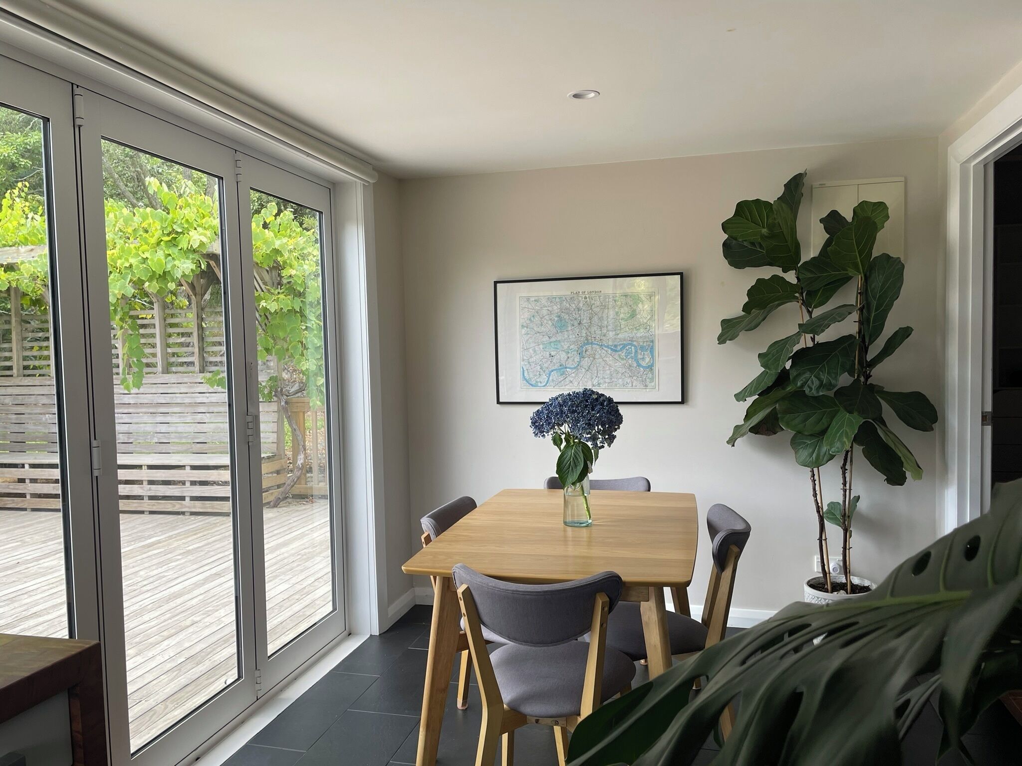 A dining area with a wooden table and chairs, a vase with flowers, a map on the wall, and a large plant.