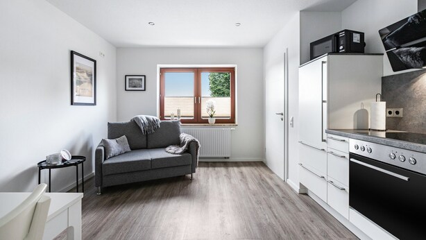 A modern kitchen with a built-in oven, a grey sofa, and a window with blinds. - Apartment '2' with Wi-Fi (Weddingstedt/OT Weddinghusen)