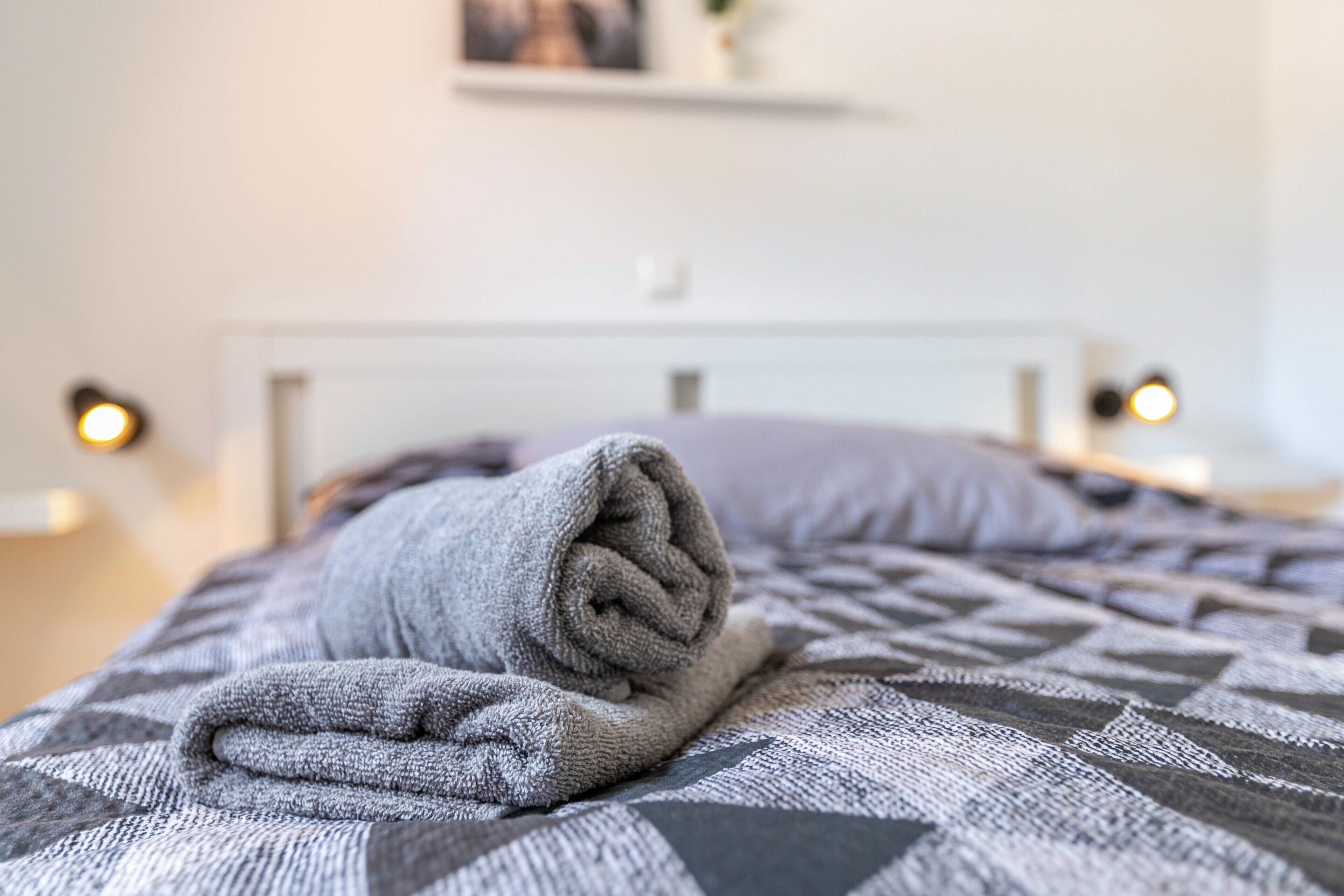 A neatly made bed with a patterned blanket and a rolled towel on it.