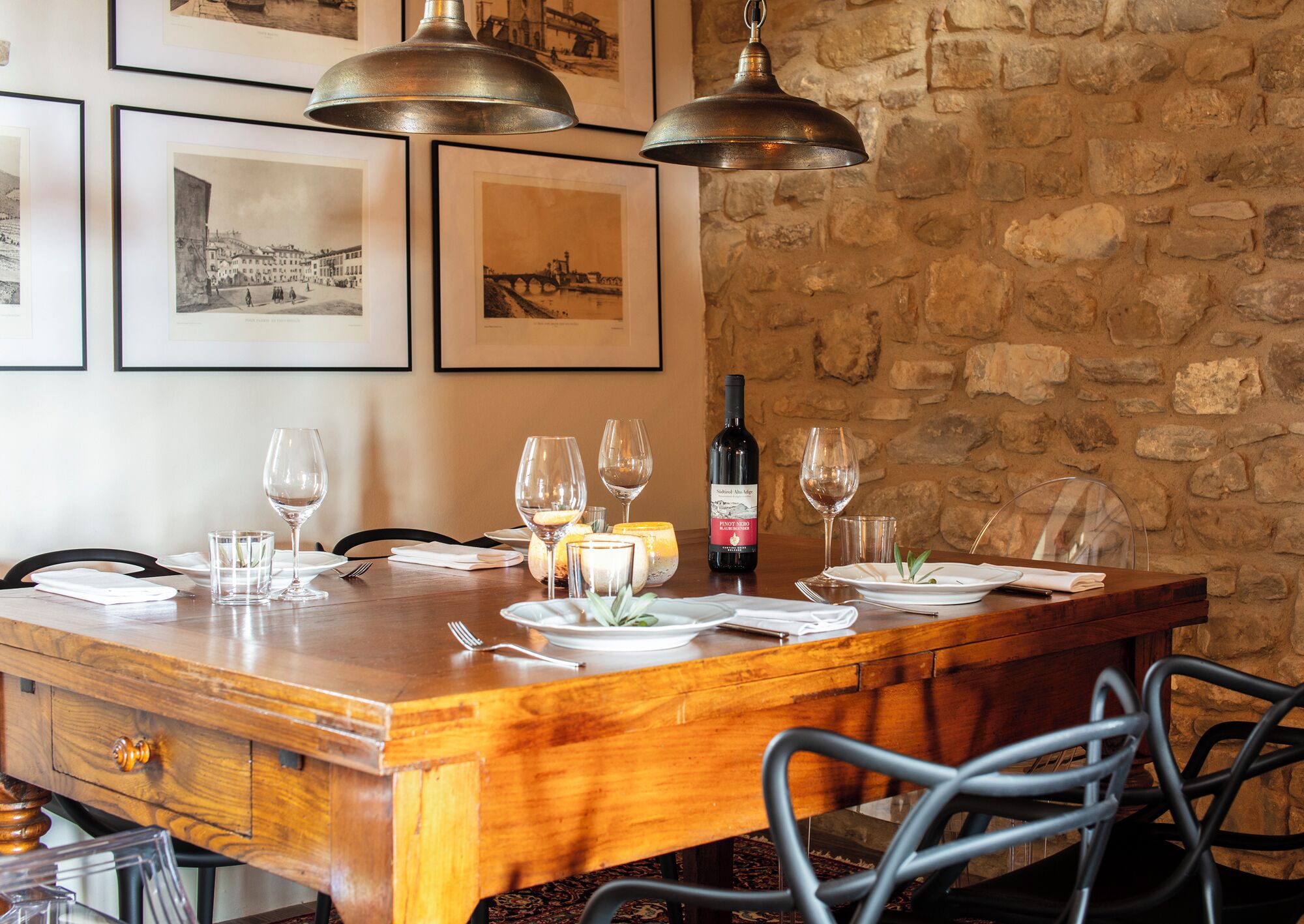 Una tavola imbandita con bicchieri da vino, piatti e una bottiglia di Pinot Nero. La tavola è circondata da sedie e appoggiata a un muro di pietra con quadri appesi.