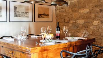 A dining table set with wine glasses, plates, and a bottle of Pinot Nero wine. The table is surrounded by chairs and set against a stone wall with framed pictures on the wall.