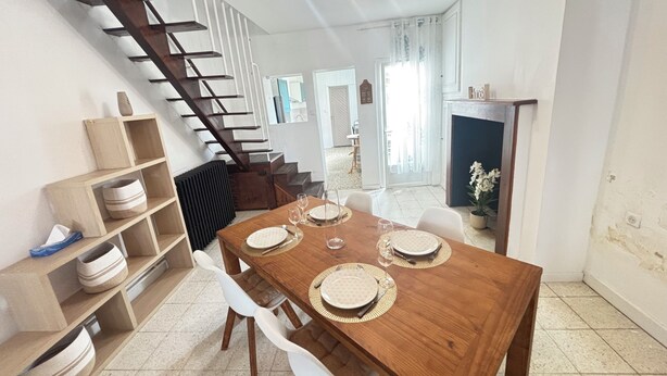 A dining area with a wooden table set for four, white chairs, a staircase, and a fireplace. - Cosy 3-bedroom house - ideal for families and groups (Brionne)