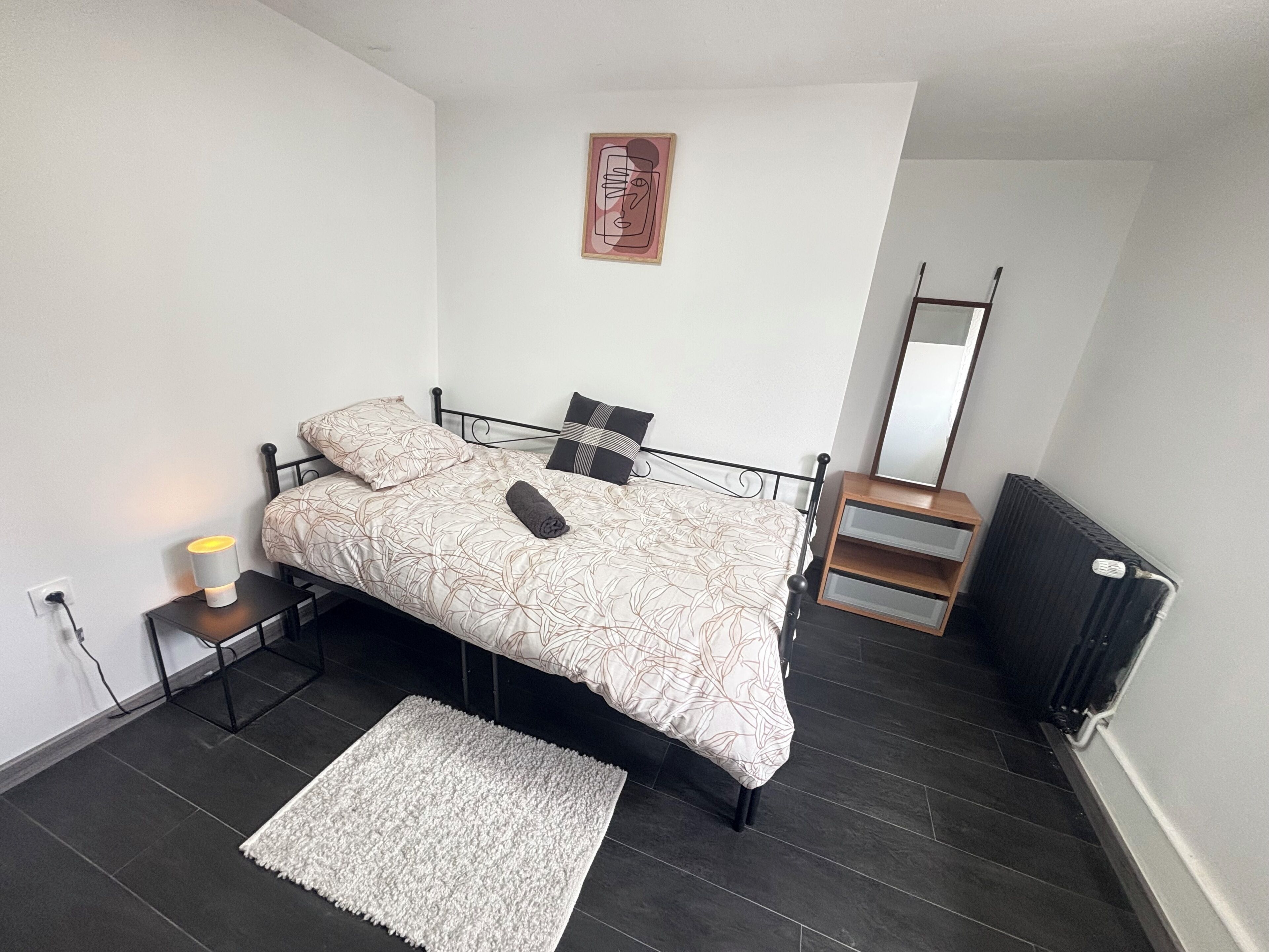 Une chambre à coucher avec un lit, une lampe de chevet, un radiateur et une petite table de nuit en bois.