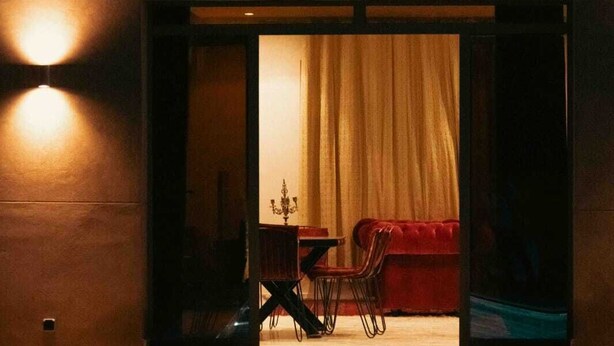 A modern interior with a red sofa and a dining table, a large window with curtains, and a pool in the foreground. - Villa Zeitouna (Ourika)