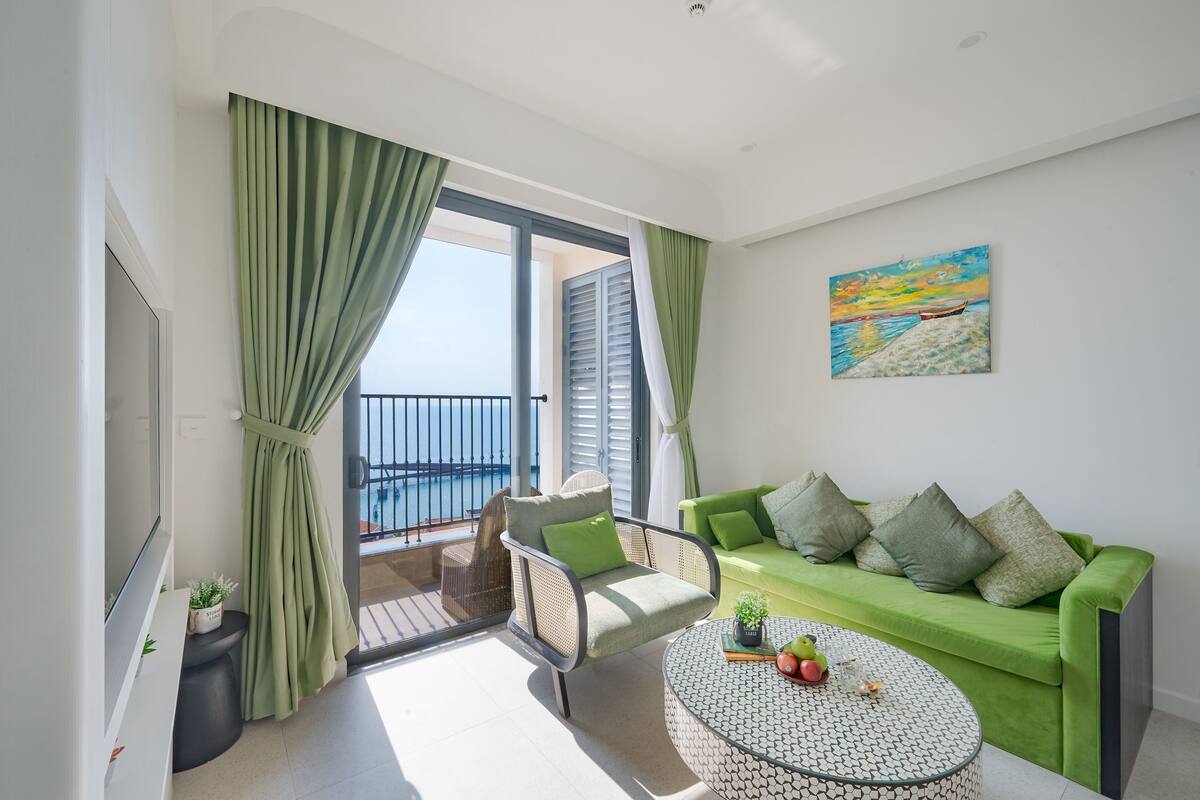 A modern living room with a green sofa, a round coffee table, and a balcony with a view of the sea.