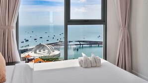A hotel room with a large window offering a view of the sea, a bridge, and a circular building. - CITY APARTMENT SUNSET TOWN (Phu Quoc)