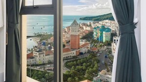 A room with a large window offering a view of a coastal cityscape, including a prominent church and colorful buildings. - CITY APARTMENT SUNSET TOWN (Phu Quoc)