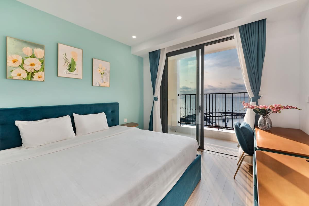 A hotel room with a large bed, a desk, and a balcony with a view of the sea.