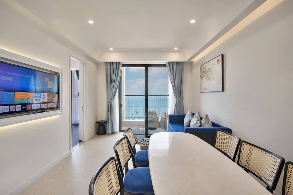 A modern hotel room with a large flat-screen TV displaying various channels, a balcony with a view of the sea, a dining area with a table and chairs, and a blue sofa with cushions.
