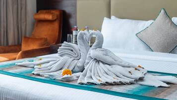 Una cama bien hecha con toallas blancas dispuestas en forma de corazón, un corredor de cama color aguamarina y un pequeño ramo de flores amarillas.