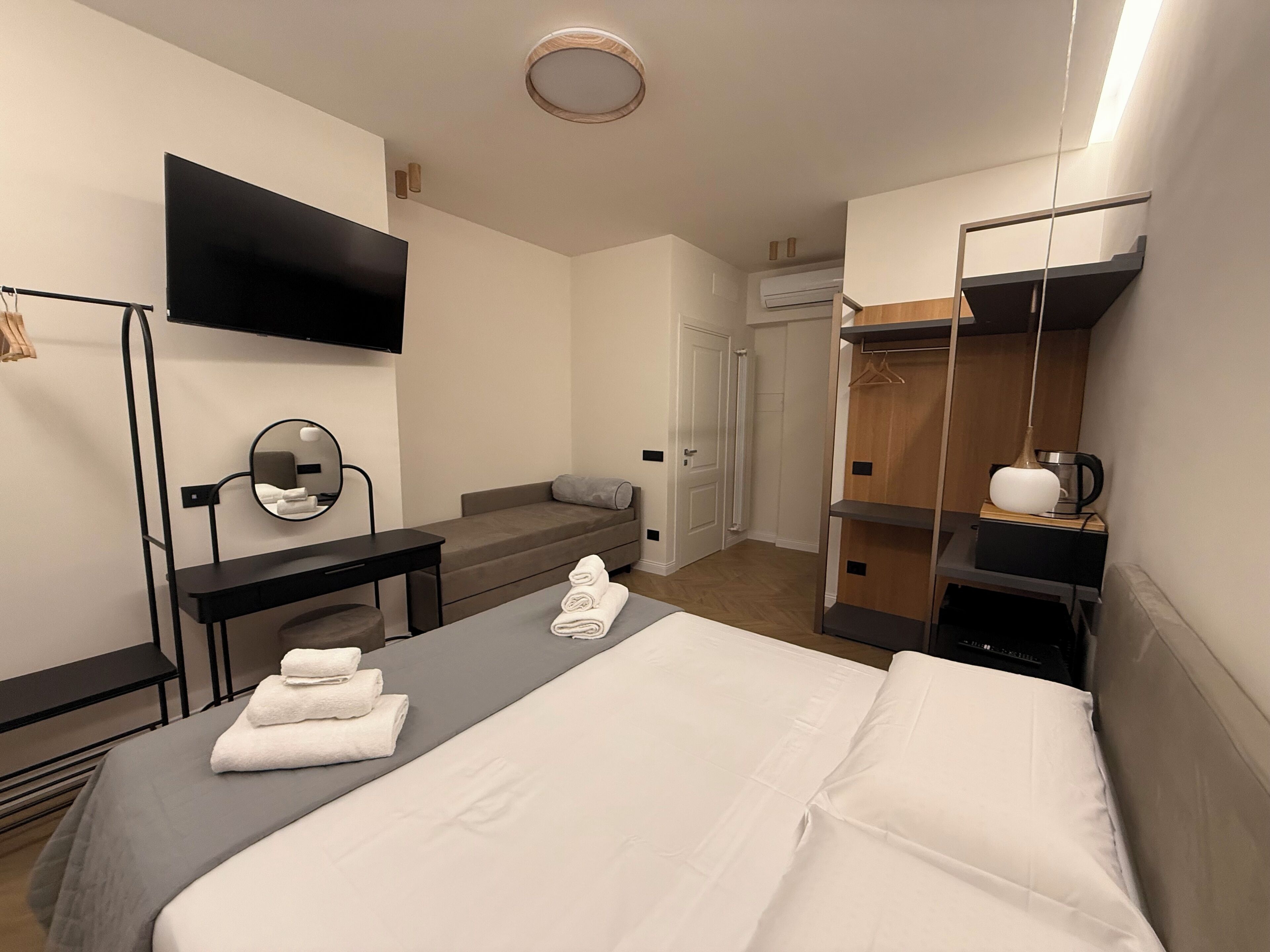 A modern hotel room with a bed, a desk, a mirror, a TV, a wardrobe, and a coffee machine.