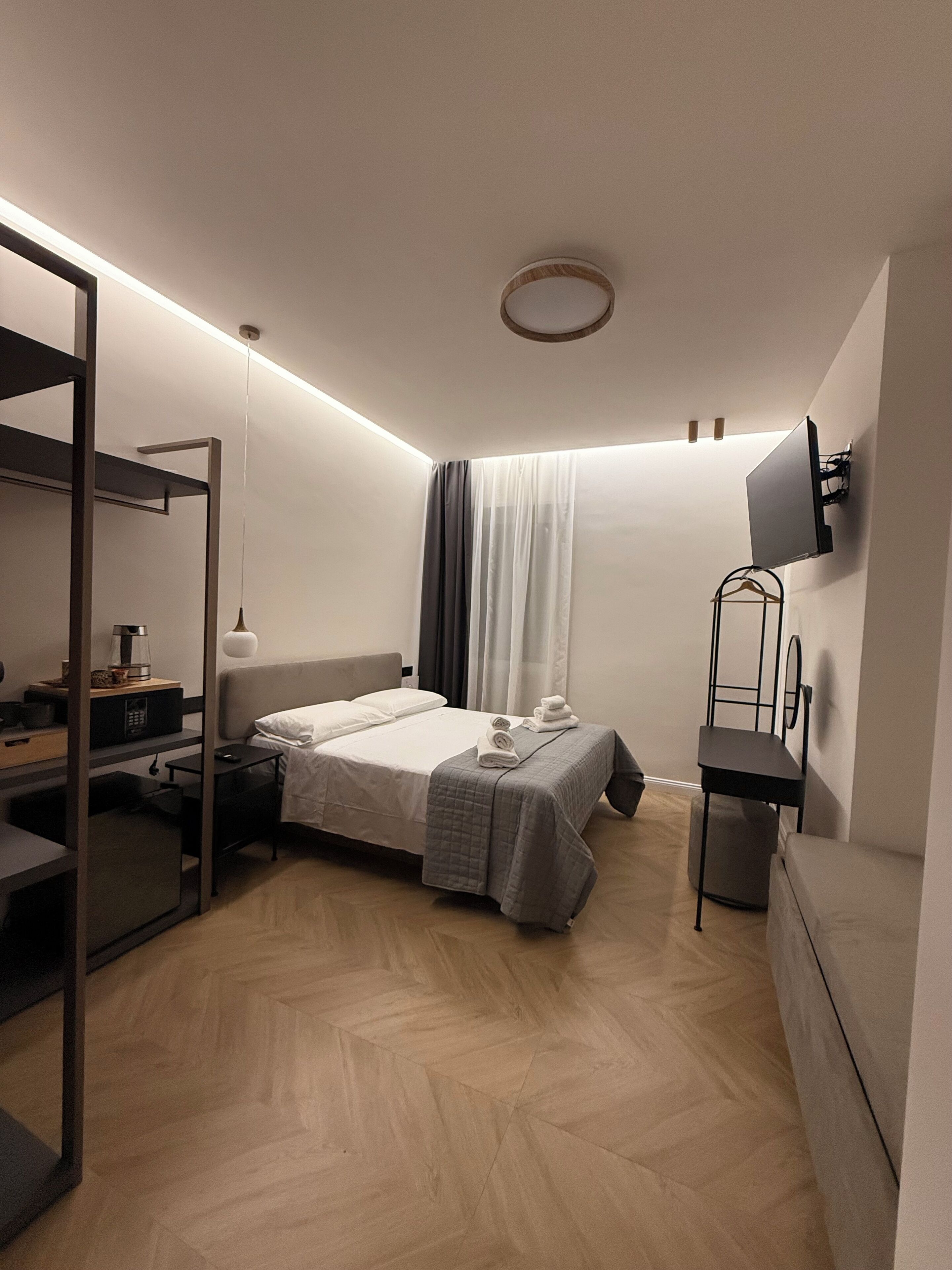 Ein kleines Hotelzimmer mit Bett, Nachttisch, Lampe und einem an der Wand befestigten Fernseher.