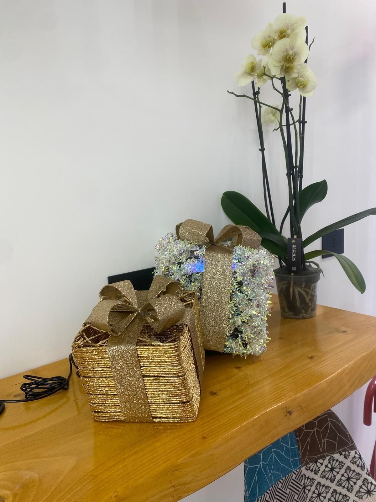 Two gift boxes on a wooden shelf, one wrapped in gold glitter and the other in burlap with a gold bow. A potted orchid plant is also visible.