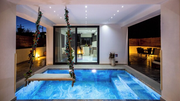 Ein modernes Hallenbad mit klarem, blauem Wasser, umgeben von Fliesenboden und -wänden. Zwei große Fenster mit Vorhängen sind zu sehen, und im Hintergrund ist ein Essbereich mit Stühlen und einem Tisch erkennbar.