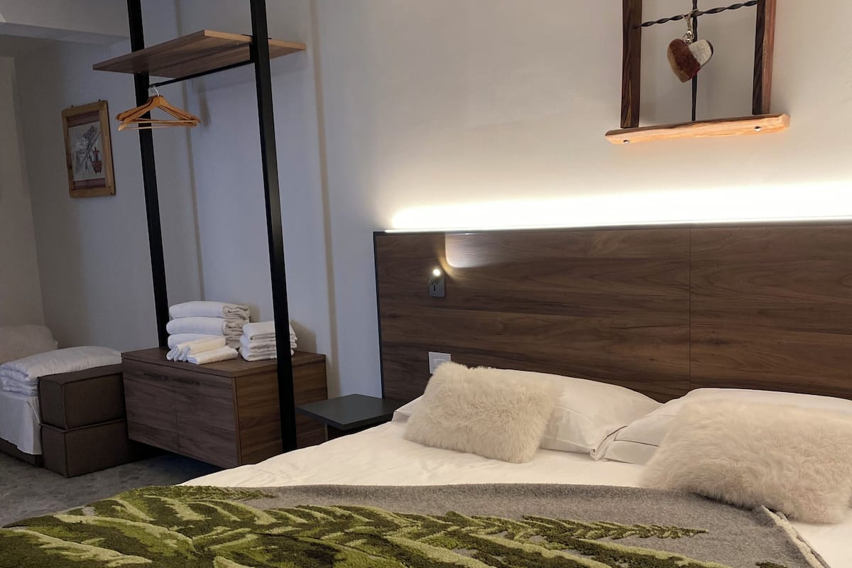 Une chambre d’hôtel moderne avec un grand lit, une tête de lit en bois, une table de chevet, un luminaire suspendu et une étagère murale où sont posées des serviettes.