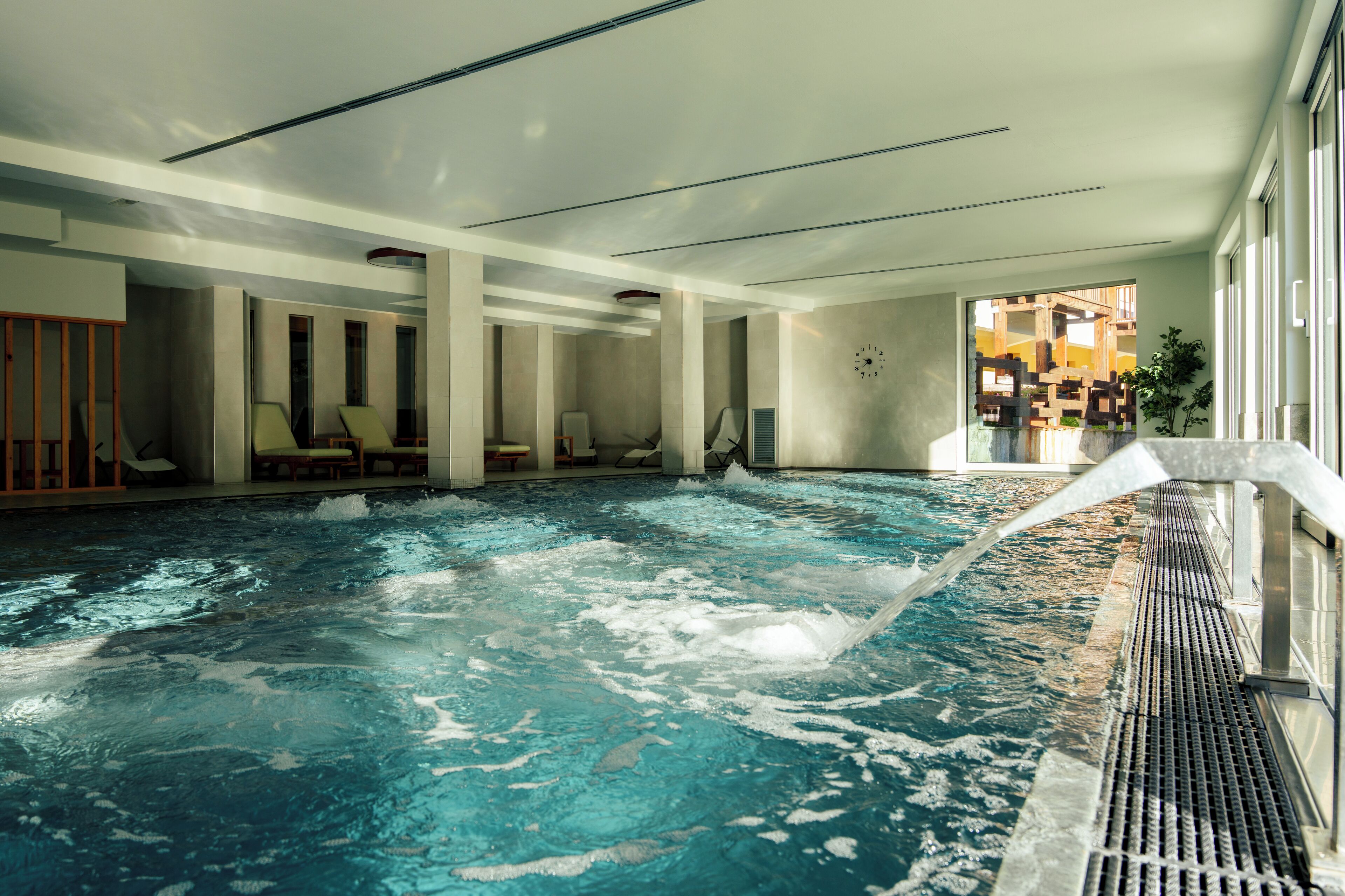Une piscine intérieure à l’eau claire, entourée de chaises longues et offrant une vue sur un bâtiment à travers un mur de verre.