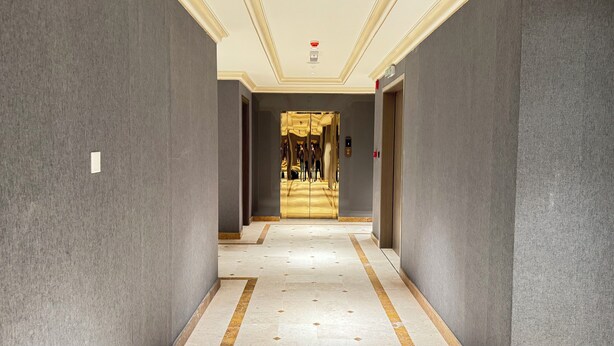 A hallway with grey walls, a marble floor, and a golden elevator door. - Amaris Hotel (Barka)