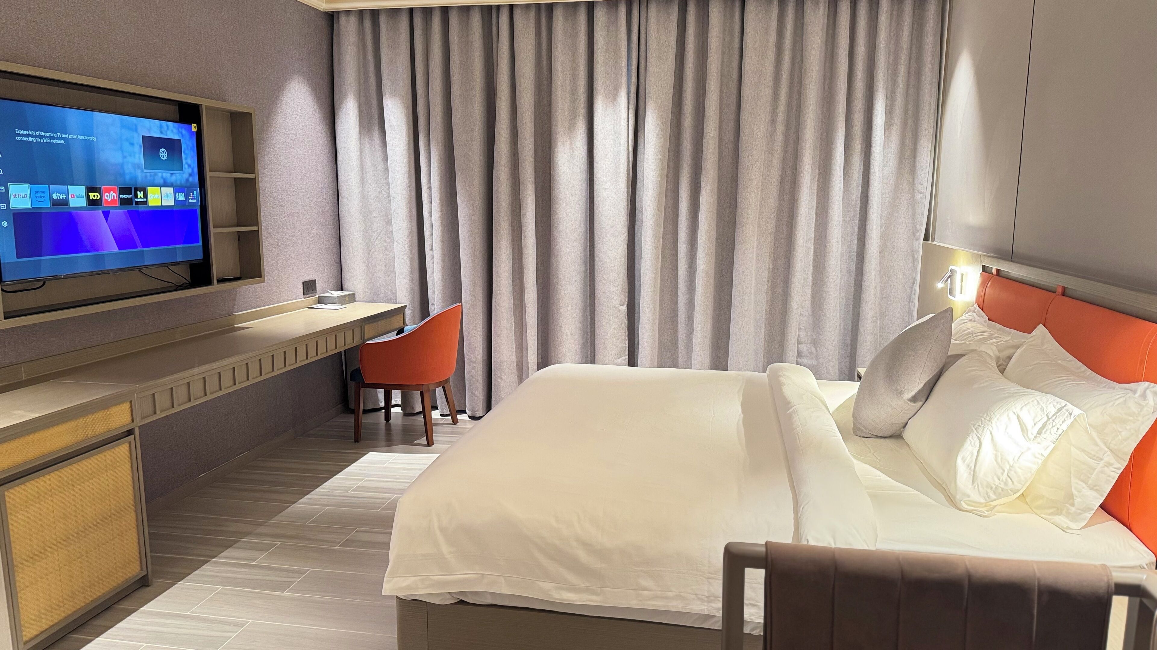Habitación de hotel con cama, escritorio, silla, televisión y un ventanal con cortinas.