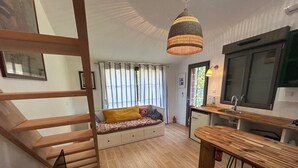 Un espace de vie compact doté d’un escalier en bois, d’un banc intégré avec des coussins, d’un coin cuisine avec un plan de travail en bois et des tabourets de bar, ainsi que d’une suspension lumineuse.