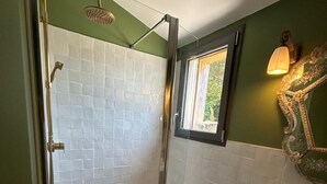 Une salle de bain dotée d’une douche à parois de verre, de murs recouverts de carreaux blancs, d’un plancher en bois, d’un lavabo blanc avec un robinet doré et d’un miroir décoratif orné d’un cadre floral.