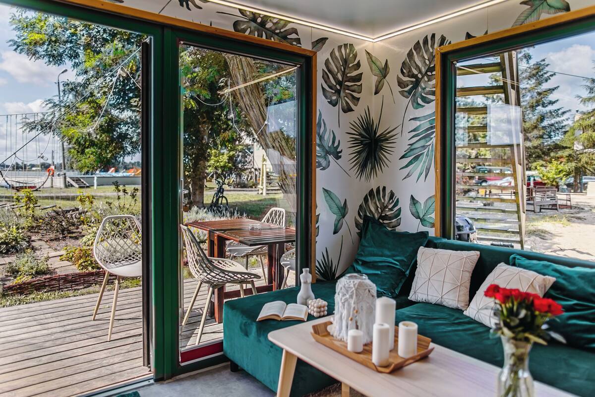 Quarto com sofá verde, mesa de madeira com velas e vista para um jardim e área externa com assentos.