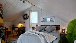Ein gemütliches Schlafzimmer mit Holzbalkendecke, einem Bett mit grauer Bettwäsche, einem Nachttisch mit Lampe und einem gerahmten Bild an der Wand.