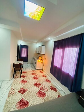 Une pièce de taille modeste comprenant un lit, un bureau, une chaise, un petit meuble de rangement, une lampe et une fenêtre avec des rideaux.