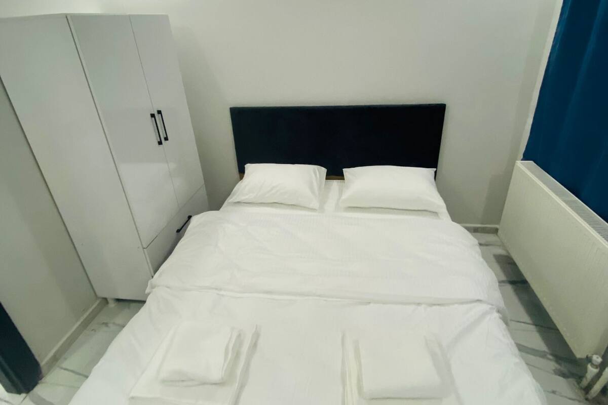 Un lit bien fait, avec du linge de lit et des serviettes blancs, une tête de lit sombre et une armoire blanche dotée d’une poignée de porte.