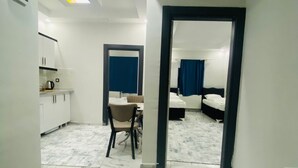 Ein Hotelzimmer mit einem Bett, einem kleinen Tisch und einem Stuhl. Es gibt eine Küchenzeile mit Schränken und einer Arbeitsfläche. Der Boden ist mit Fliesen belegt und die Decke wird von einer Leuchte erhellt.