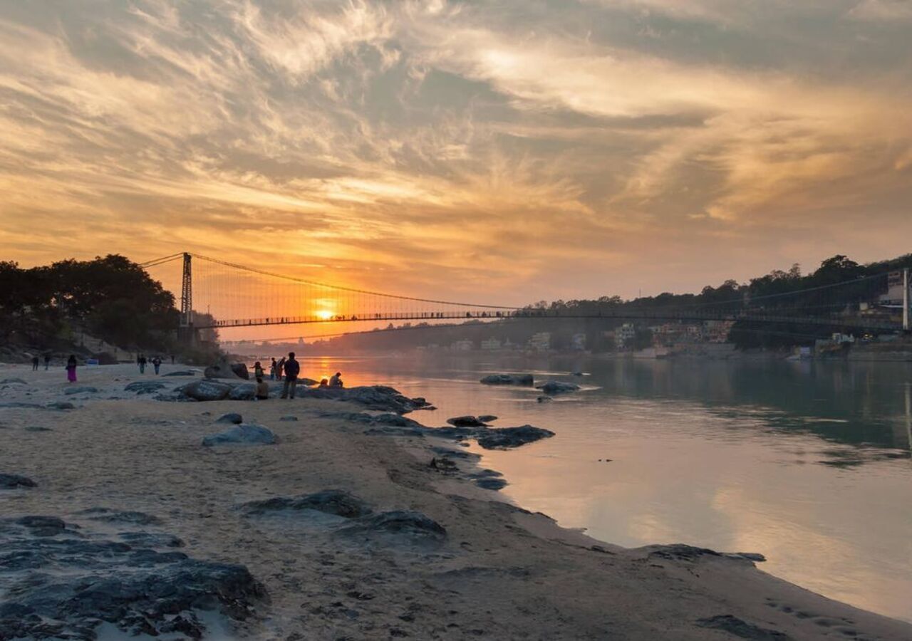 Een rivier bij zonsondergang, met een brug, een rotsachtige oever en mensen op het strand.