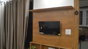 A living room with a flat-screen TV mounted on a wooden wall, a power outlet, and a shelf with a plant. - AniGanges - Quiet 1-Bedroom AC Retreat in Veerbhadra, Rishikesh (Veerbhadra)