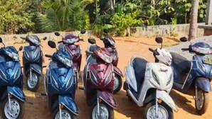 A row of scooters parked on a dirt path with greenery in the background.