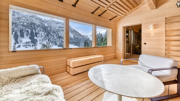 Interior de una cabaña de madera con un gran ventanal que da a un paisaje montañoso nevado, una mesa redonda y un sofá.