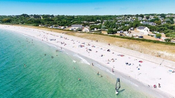 Vista aérea de una playa con agua turquesa cristalina, una orilla arenosa y edificios al fondo.