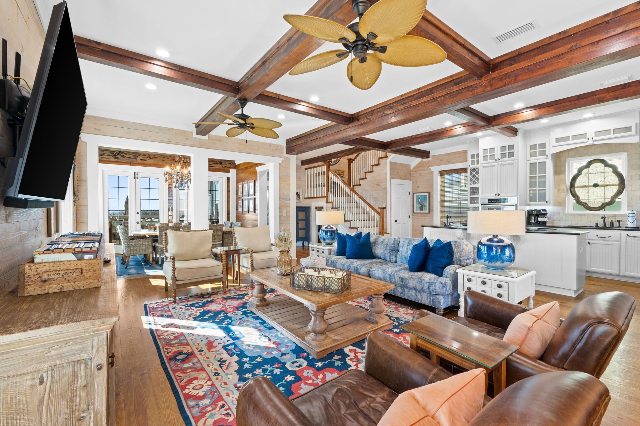 A spacious living room with a large wooden ceiling, a fireplace, a television, and a kitchen area with white cabinets and a large window.