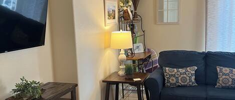Ein Wohnzimmer mit Flachbildfernseher, einem dunklen Holztisch, einem blauen Sofa mit gemusterten Kissen, einem kleinen Beistelltisch mit Lampe und einer Zierpflanze.