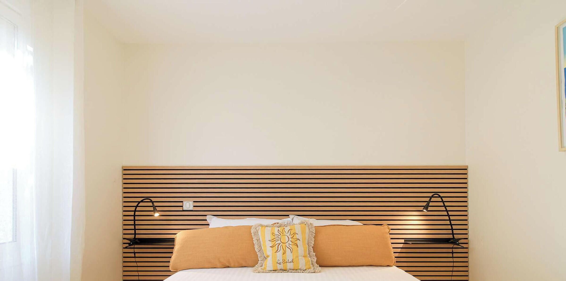 Une chambre à coucher avec une tête de lit en lattes de bois, un lit avec une literie blanche et deux oreillers, dont l’un est orné d’un motif solaire.